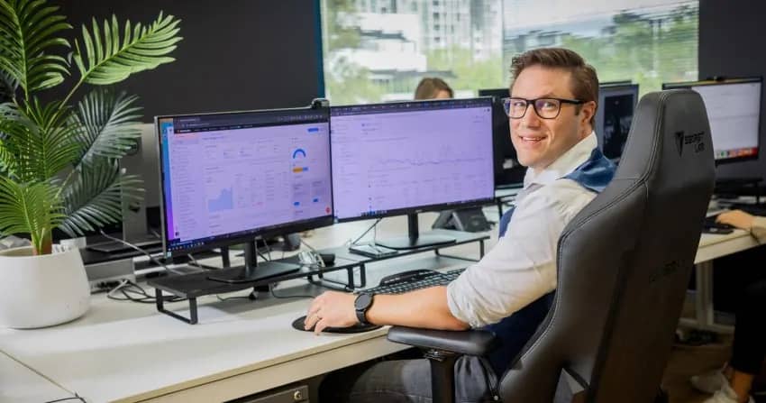 team member sitting at a dual-monitor workstation with semrush analytics dashboards open on screen, wearing glasses and a white shirt, with a potted plant and natural light from the office windows.