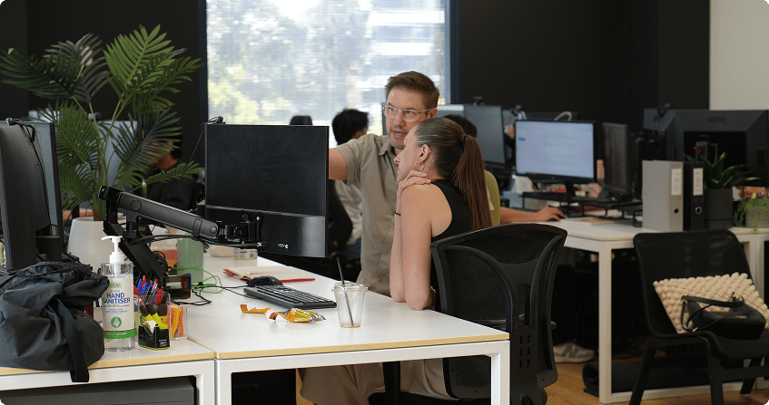 two team members collaborating at a workstation in an open-plan office, one standing and pointing at the screen while the other is seated, with multiple monitors and indoor plants visible in the background.