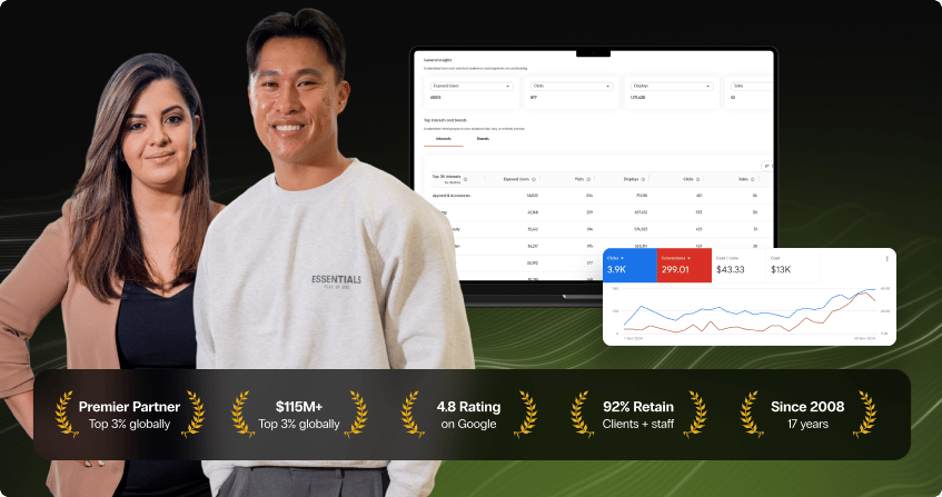 two team members at a white desk in a dark-walled office, one working on a desktop computer while the other reviews data on a laptop, with a potted plant and coffee mug on the desk.