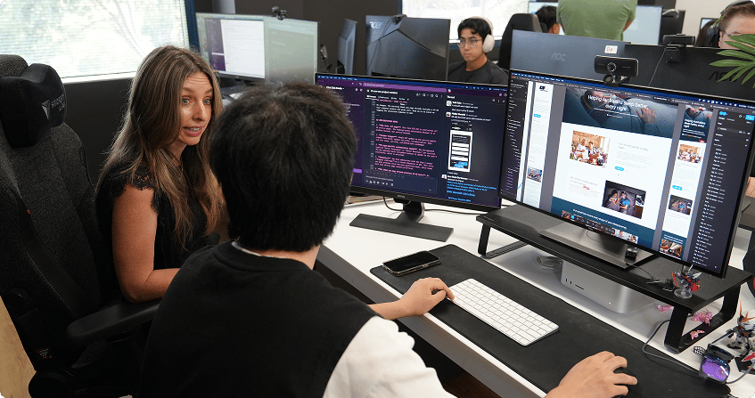 web designer leaning over to discuss a project with a developer at a dual-monitor workstation showing slack messages and a live website build in the browser.