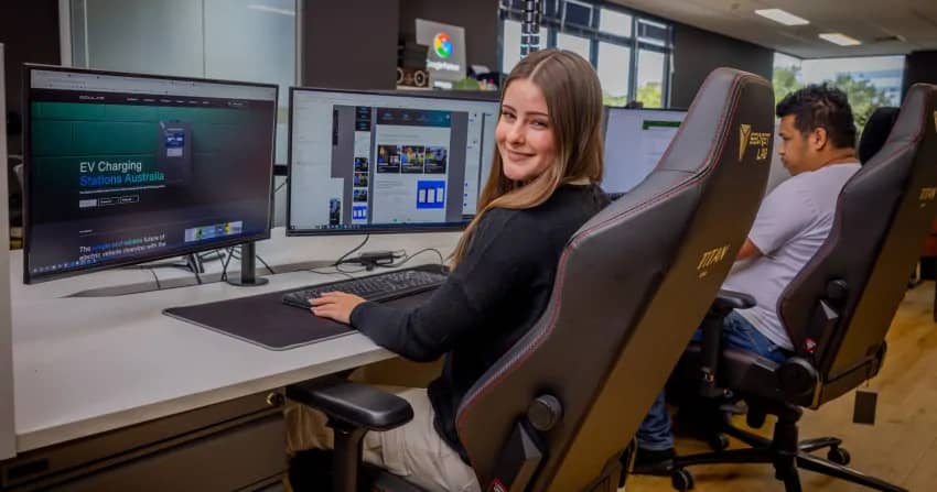 female developer sitting at a triple-monitor workstation with an ev charging stations australia website visible on screen, smiling at the camera with a colour wheel app open on a secondary display, in the open-plan norwest office with another team member working behind her.