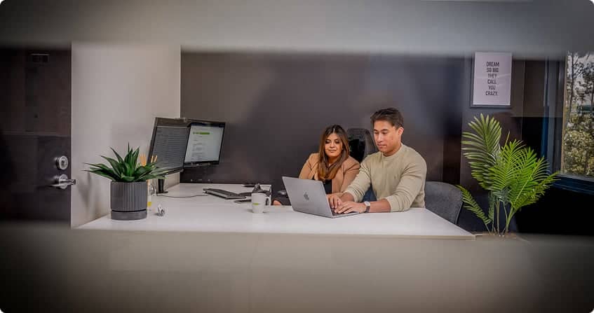 two google ads specialists sitting side by side at a white desk reviewing campaign data on a laptop in a modern office with dark walls and natural light.