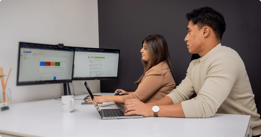 
two team members at a white desk with dual monitors showing campaign data and ad management interfaces, one working on a laptop while the other reviews the screens.