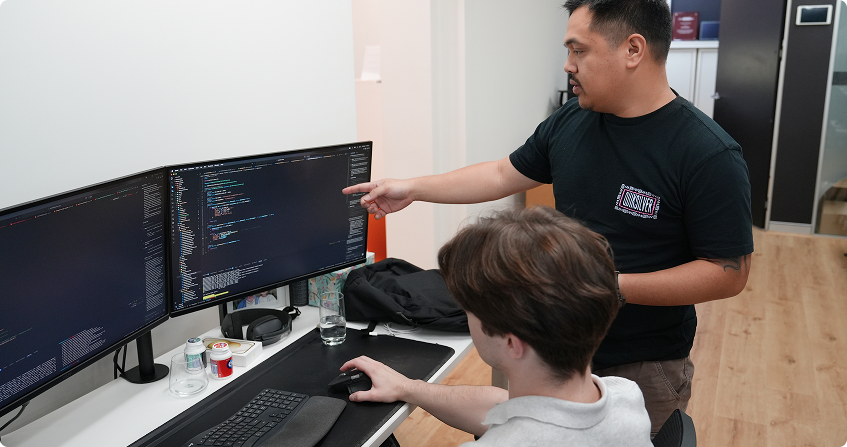 two paladine systems developers at a triple-monitor workstation reviewing code together, with one standing and pointing at the screen while the other navigates.