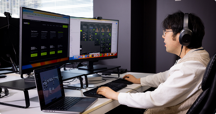 young man wearing headphones working across a triple-monitor setup with a pricing page design on the main screen, a project management tool on the second, and a laptop showing a wireframe below.