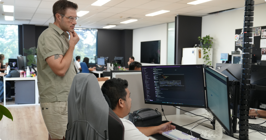 team member standing and reviewing code on a dual-monitor setup while another team member works at the keyboard, in an open-plan office with multiple workstations and natural light from large windows.