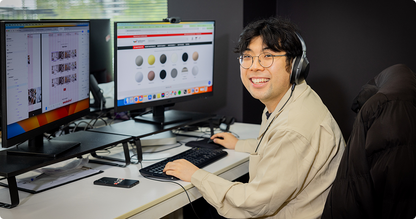 jerry smiling at the camera from his dual-monitor workstation wearing headphones, with an ecommerce product colour swatch page open on one screen and a product image gallery on the other.