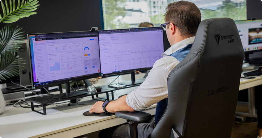team member sitting at a dual-monitor workstation reviewing analytics dashboards and audit data, wearing a white shirt and glasses, with a potted palm and natural light from office windows.