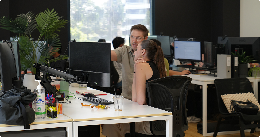 two team members collaborating at a workstation in an open-plan office, one standing and pointing at the screen while the other is seated, with multiple monitors and indoor plants visible in the background.