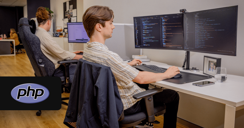 developer sitting at a dual-monitor workstation with php code visible on both screens, with the php logo overlaid, in the open-plan norwest office with another team member working behind him.