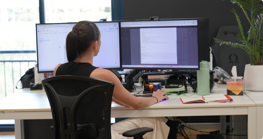 team member working at a dual-monitor desk reviewing website data and content, with a red notebook open beside the keyboard, in a bright office with natural light.