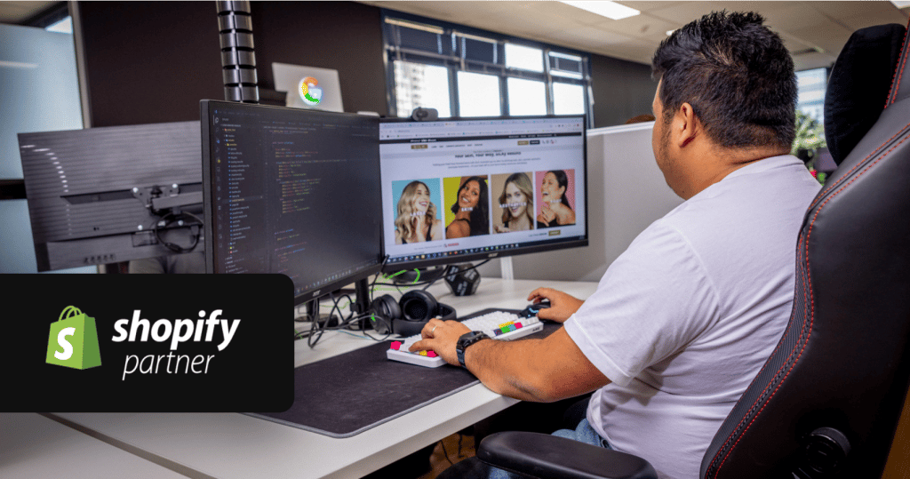developer sitting at a workstation reviewing shopify code on a dual-monitor setup, with a design mockup and product photography visible on the secondary screen, in the open-plan norwest office.
