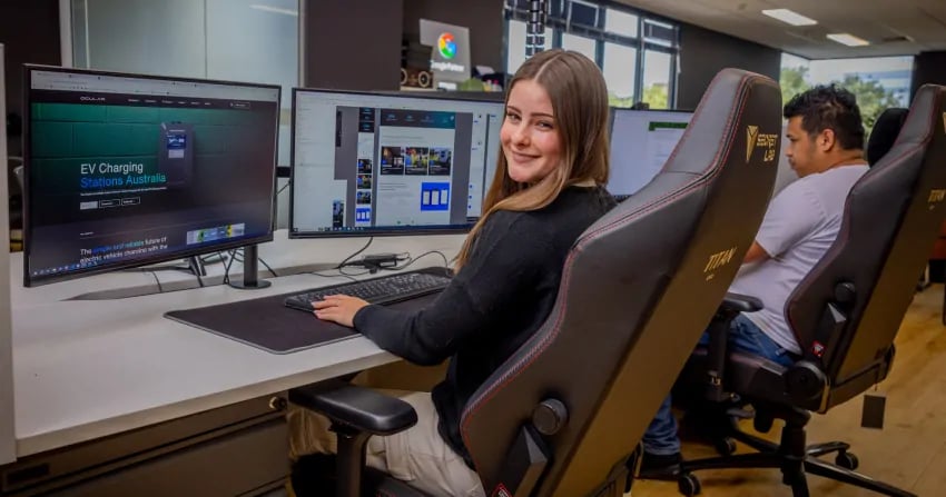 female developer sitting at a triple-monitor workstation with an ev charging stations australia website visible on screen, smiling at the camera with a colour wheel app open on a secondary display, in the open-plan norwest office with another team member working behind her.