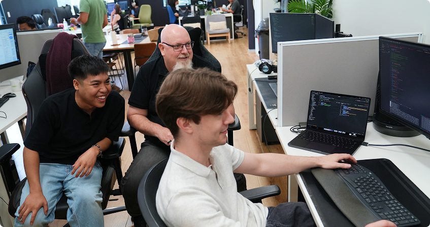 three click click media team members gathered around a workstation reviewing code on a laptop and monitor, laughing while collaborating in the open-plan norwest office.