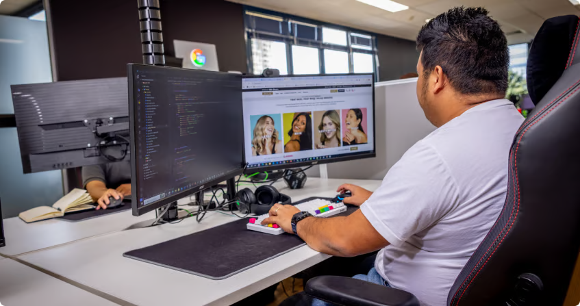 a click click media developer working at a dual-monitor workstation with code on one screen and a live ecommerce website on the other, with a colourful mechanical keyboard.