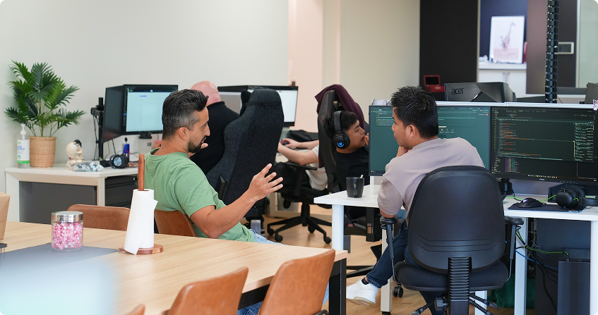 two developers having a conversation at a shared desk with code visible on dual monitors, while other team members work at their stations in the background of the open-plan norwest office.