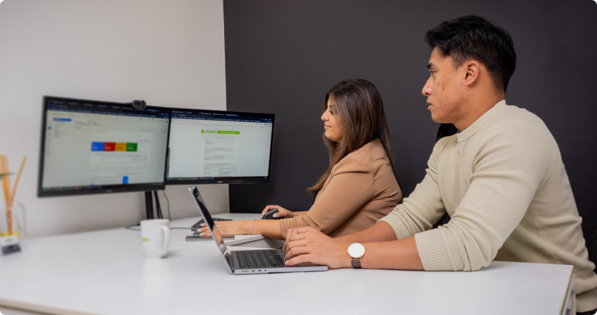 two team members at a white desk with dual monitors showing campaign data and ad management interfaces, one working on a laptop while the other reviews the screens.