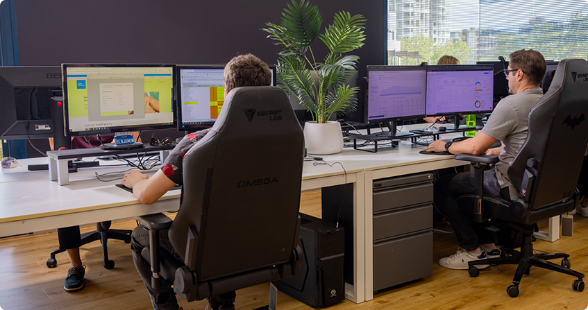 two team members working at desks with dual monitors showing analytics dashboards and content tools, with a potted palm between the workstations in a modern office with timber flooring.