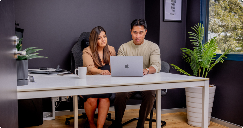 two ppc specialists sitting side by side at a white desk reviewing campaign data on a laptop in a modern office with dark walls, a framed quote, and a potted plant.